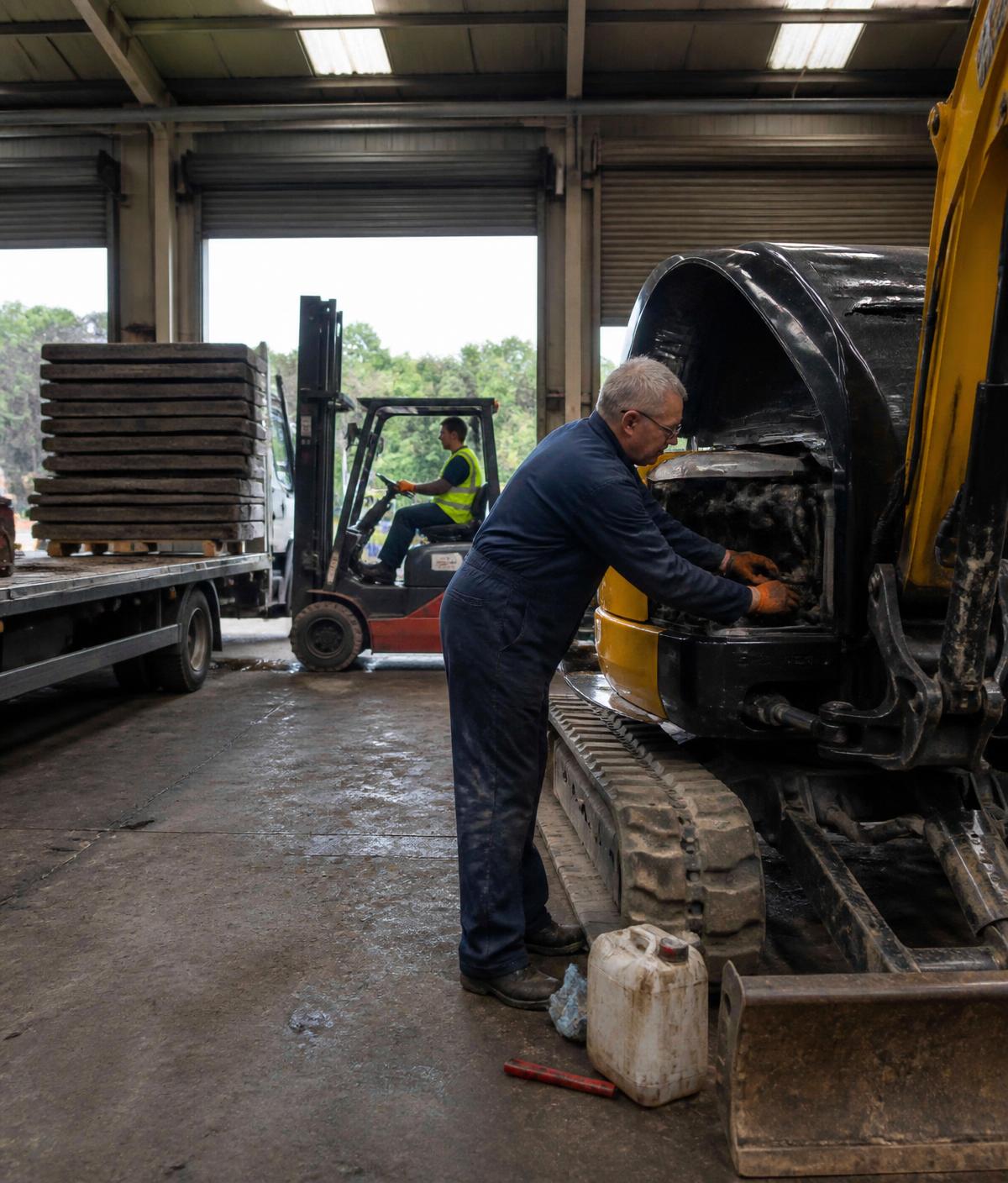 NLPH engineer servicing a mini excavator while a colleague loads ground protection mats at the depot